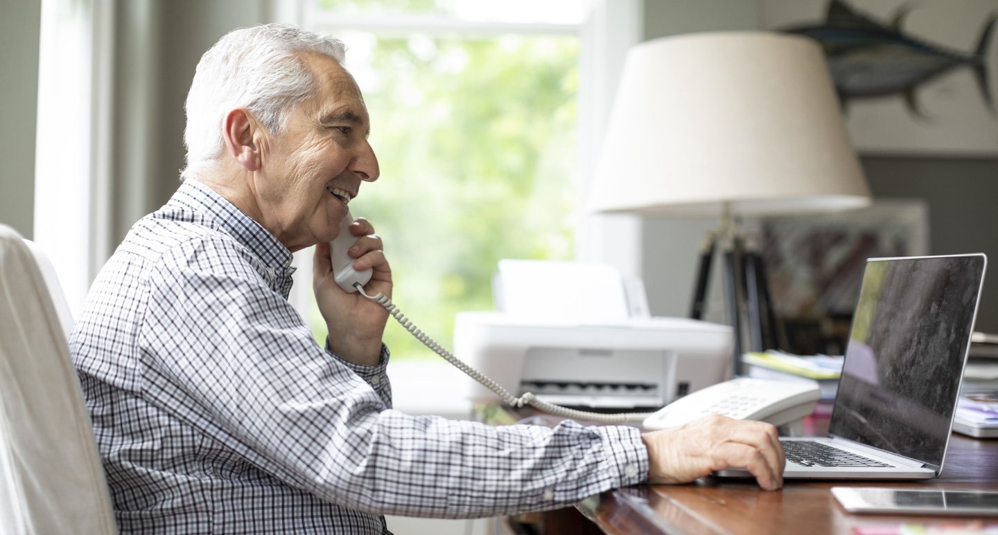 Äldre man pratar i telefon och arbetar med dator och skrivare