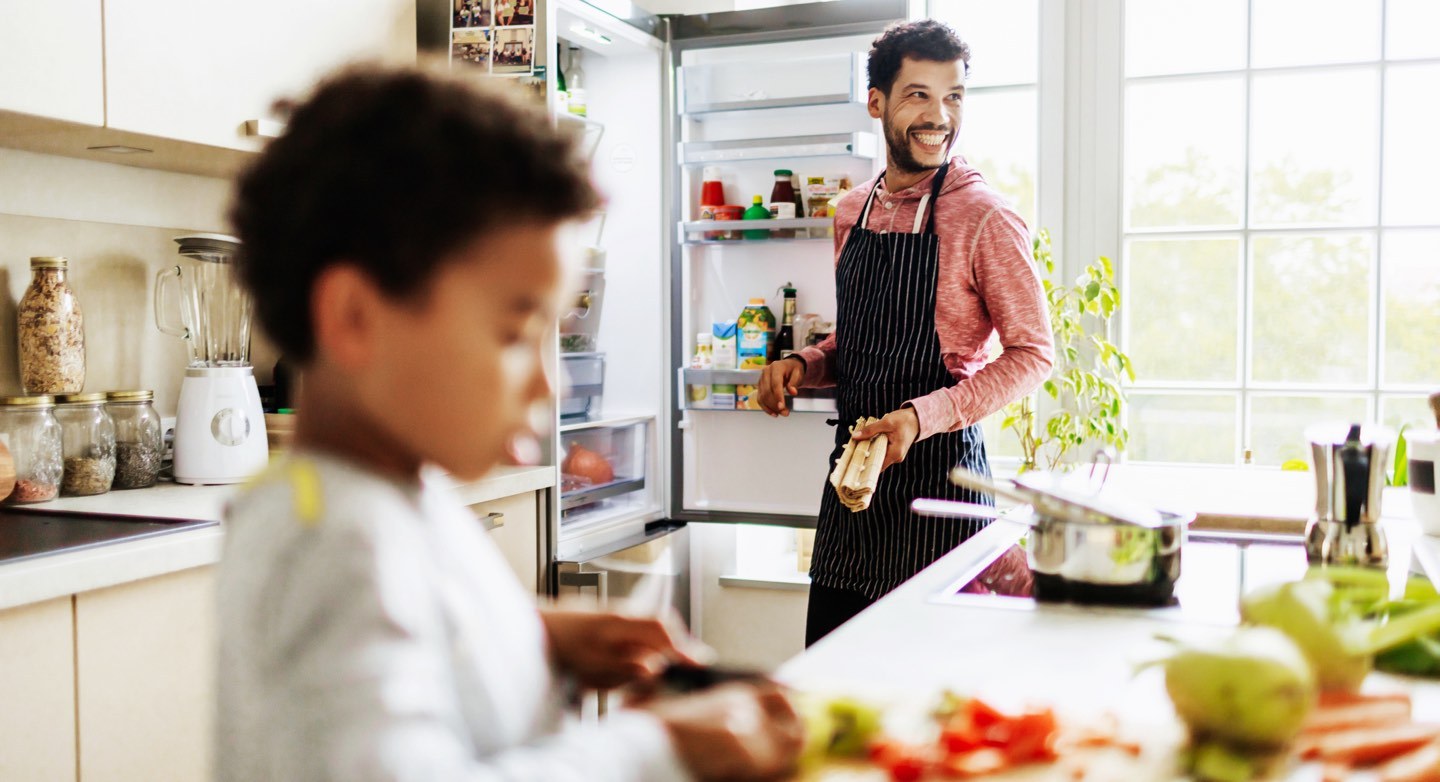 Glad man vid ett öppet kylskåp, och ett barn som skär grönsaker